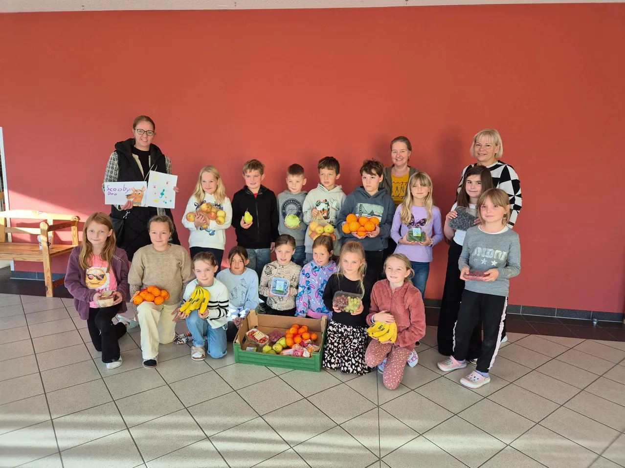 Eine Gruppe von Kindern und Erwachsenen posiert für ein Foto mit verschiedenen Obst- und Gemüsesorten. Zwei Frauen stehen hinter ihnen.
