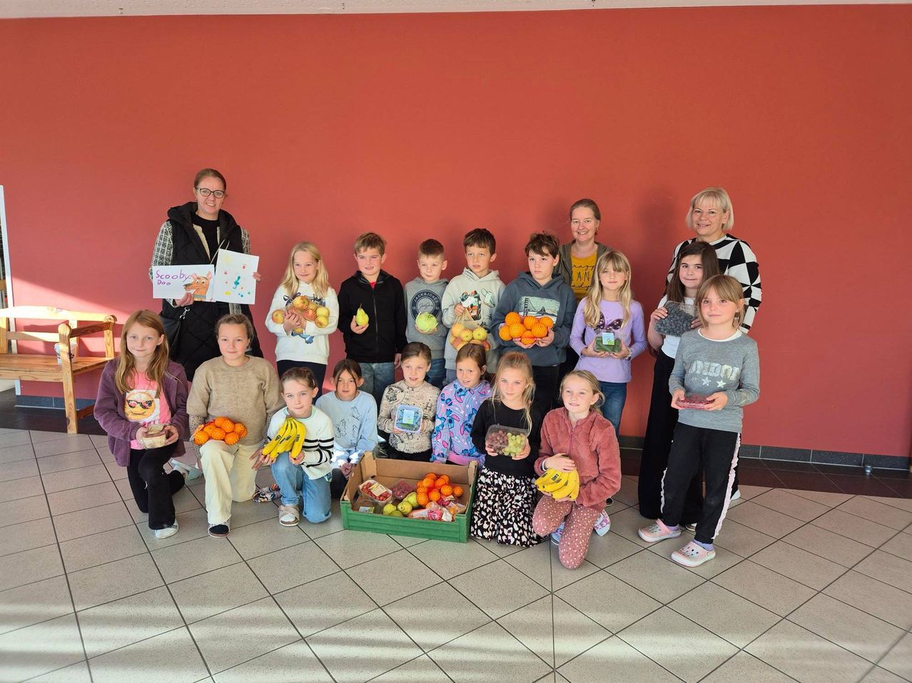 Eine Gruppe von Kindern und Erwachsenen posiert für ein Foto mit verschiedenen Obst- und Gemüsesorten. Zwei Frauen stehen hinter ihnen.