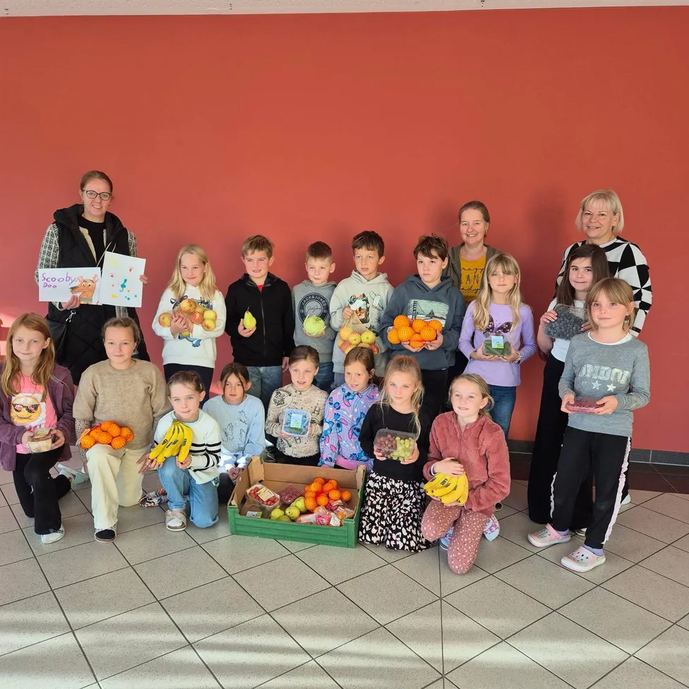 Eine Gruppe von Kindern und Erwachsenen posiert für ein Foto mit verschiedenen Obst- und Gemüsesorten. Zwei Frauen stehen hinter ihnen.