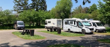 Mehrere Wohnmobile sind in einem bewaldeten Gebiet geparkt, mit Bäumen und einem klaren blauen Himmel im Hintergrund. Es gibt Picknicktische und Mülleimer in der Nähe.