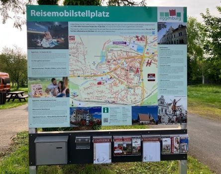 Ein Wegweiser zeigt eine Karte einer Stadt mit verschiedenen Sehenswürdigkeiten und einem Schild für Reisemobilparkplätze in Grün und Weiß. Unter der Karte befinden sich Broschüren und Postkarten, und in der Nähe steht eine Picknickbank.