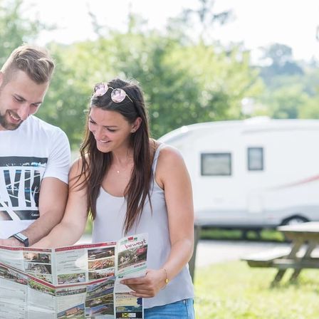 Ein Paar steht in einem Park mit einem Reiseführer, lächelt und schaut sich den Inhalt an. Hinter ihnen steht ein Wohnmobil, und ein Picknicktisch ist in der Nähe.