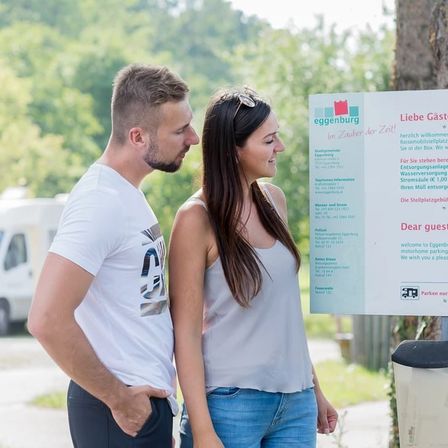 Ein Mann und eine Frau stehen vor einem Schild mit Informationen über Egensburg und schauen darauf. Der Mann hat einen Bart und ein weißes T-Shirt mit Logo, und die Frau hat lange Haare und ein weißes ärmelloses Oberteil. Sie befinden sich in einer Außenumgebung mit einem weißen Van und Bäumen im Hintergrund.