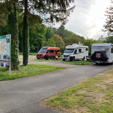 Drei Wohnmobile sind am Straßenrand in einem Grasbereich mit Bäumen geparkt. Ein Kartenschild steht links.