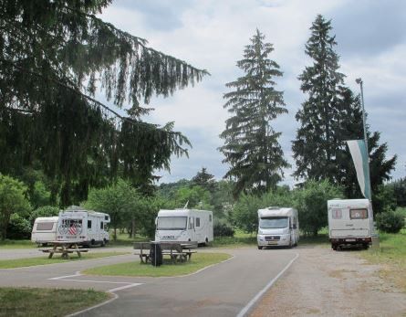 Einige Wohnmobile sind in einem Waldgebiet geparkt, mit Picknicktischen und Bänken in der Nähe. Hohe Bäume und Sträucher umgeben das Gebiet, und eine Fahne ist auf der rechten Seite sichtbar.