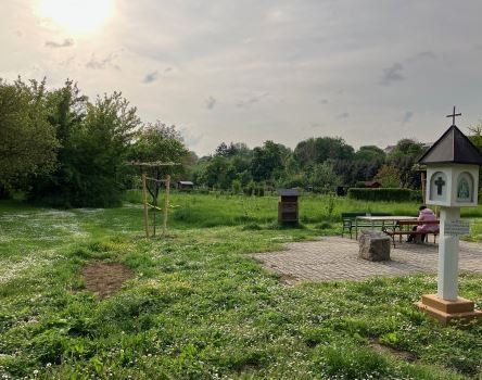 Eine friedliche Parkszene mit einer kleinen Kapelle, einer Bank und einem Picknicktisch. Bäume und Büsche umgeben das Gebiet. Eine Person sitzt auf der Bank. Der Himmel ist klar mit einigen Wolken.