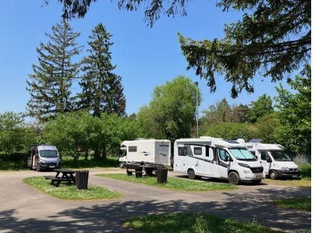 Mehrere weiße Wohnmobile sind in einem Campingplatz mit üppigen grünen Bäumen, Picknicktischen und Mülleimern geparkt. Ein Van ist ebenfalls in der Nähe geparkt.