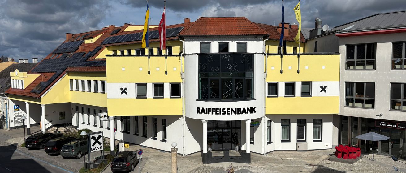 Ein Bankgebäude mit rotem Dach, gelben Wänden und Fahnen über dem Eingang. Solarmodule sind auf dem Dach installiert. Der Eingangsbereich zeigt ein Schild mit der Aufschrift Raiffeisenbank.