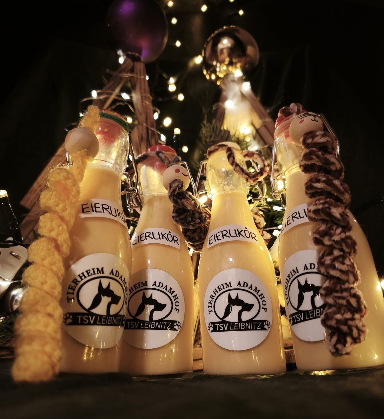 Fünf Flaschen Eierlikör mit braunen Flaschenverschlüssen und Weihnachtsdekorationen dahinter. Jede Flasche hat ein Etikett mit dem Text 'Tierheim Adamhof' und 'TSV LEIBNITZ'.