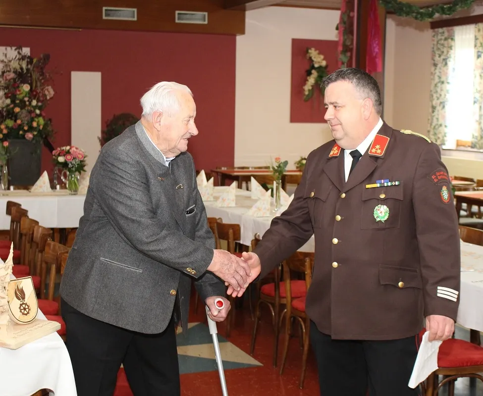Ein älterer Mann mit einem Gehstock schüttelt einem Mann in Uniform in einem dekorierten Raum die Hand.