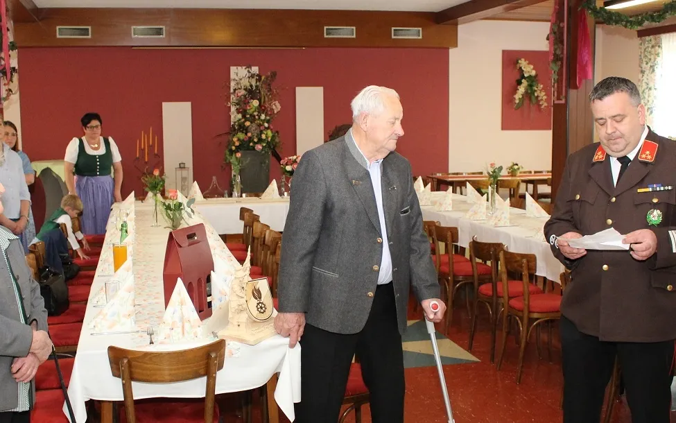 Ein älterer Mann benutzt einen Stock, um durch einen Speisesaal zu gehen, der für eine formelle Veranstaltung vorbereitet ist. Der Tisch ist mit Blumen, Servietten und Weingläsern geschmückt. Ein anderer Mann steht in der Nähe und liest ein Stück Papier.