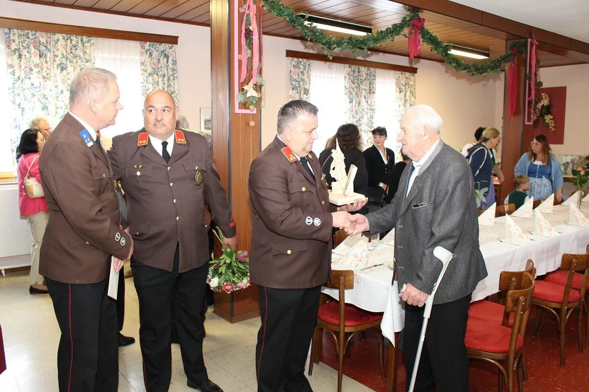 Zwei Männer in Militäruniformen schütteln sich die Hände mit einem älteren Mann, der einen Stock hält, wahrscheinlich bei einer Feier. Sie stehen in der Nähe eines Tisches mit weißer Tischdecke, umgeben von anderen.