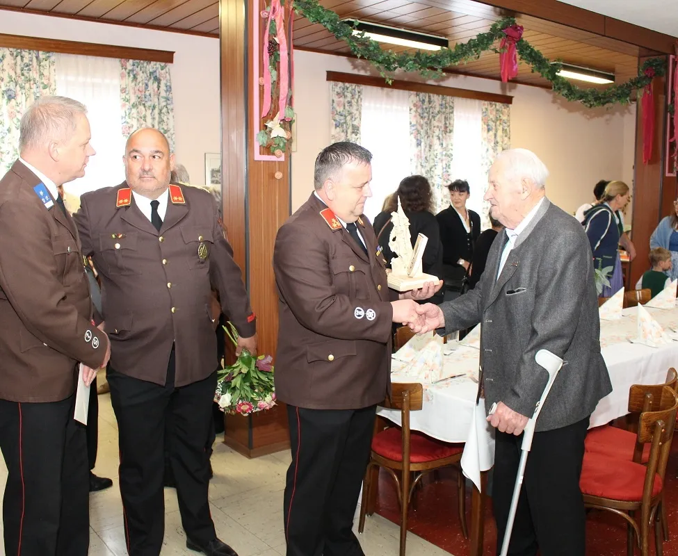 Zwei Männer in Militäruniformen schütteln sich die Hände mit einem älteren Mann, der einen Stock hält, wahrscheinlich bei einer Feier. Sie stehen in der Nähe eines Tisches mit weißer Tischdecke, umgeben von anderen.