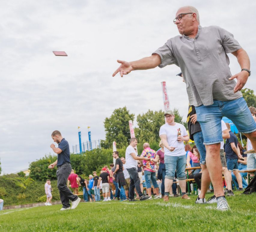 Eine Outdoor-Versammlung mit Menschen, die Frisbee auf einem grünen Feld unter einem bewölkten Himmel spielen. Bäume und ein Spielplatz sind im Hintergrund.