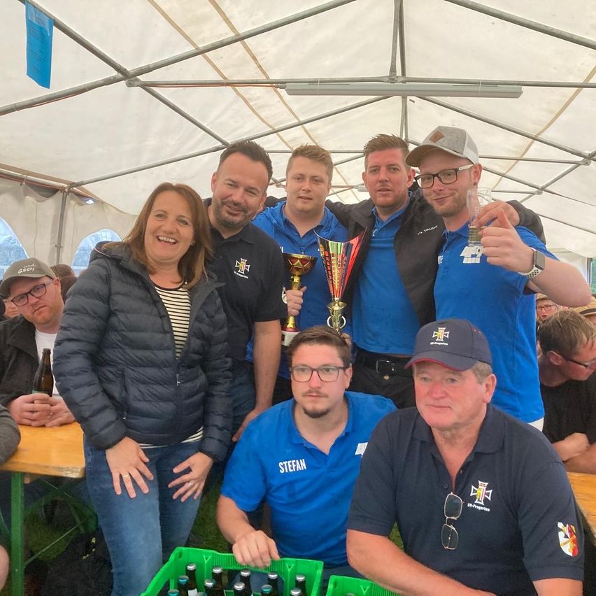 Gruppenfoto lächelnder Menschen unter einem Zelt. Sie halten Pokale und Gläser, und zwei Männer tragen Hüte. Ein Tisch hält Flaschen und Kisten Bier.