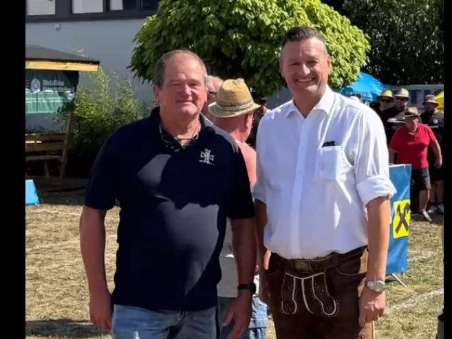 Two men stand side by side, smiling, with a tree and a blue banner with a yellow X in the background.