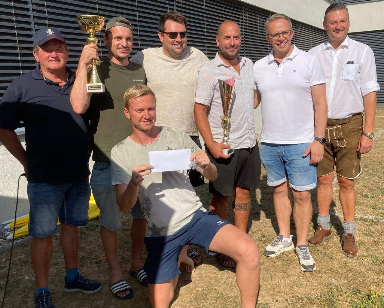 A group of men stand together, smiling, with two of them holding trophies and one holding a piece of paper. They are dressed casually and appear to be celebrating an achievement.