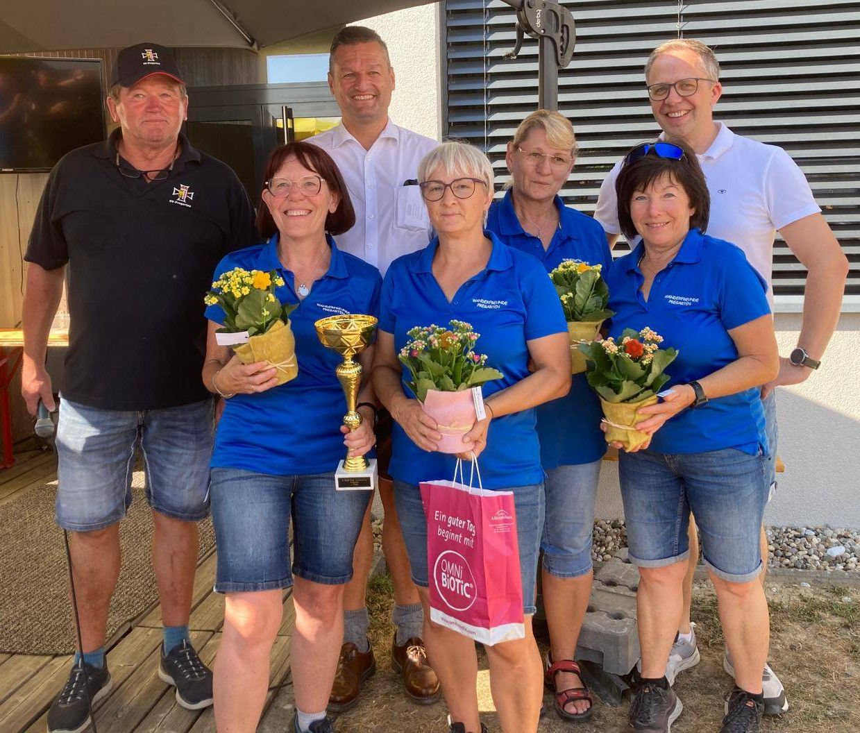 A group of people dressed in blue shirts and shorts are standing together, smiling, and holding flowers and a trophy. They appear to be celebrating a victory.