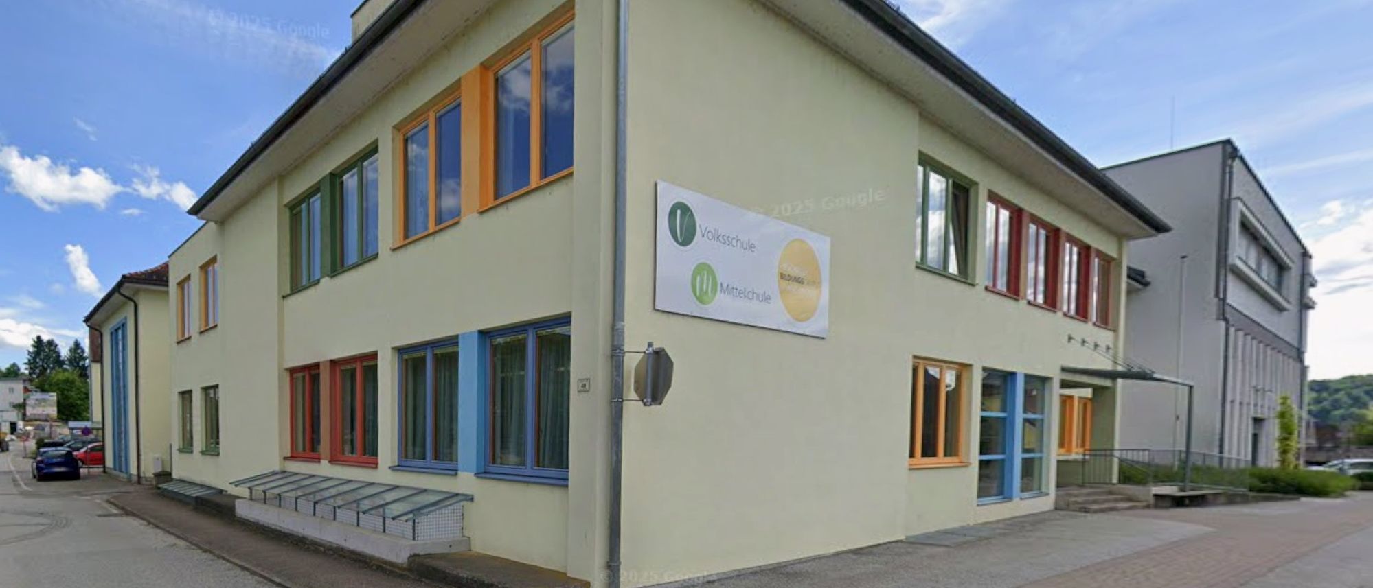 Die Vorderseite eines Gebäudes mit weißer Wand und orangefarbenen Fenstern. Es gibt ein Schild mit der Aufschrift 'Volksschule Mittelschule'.