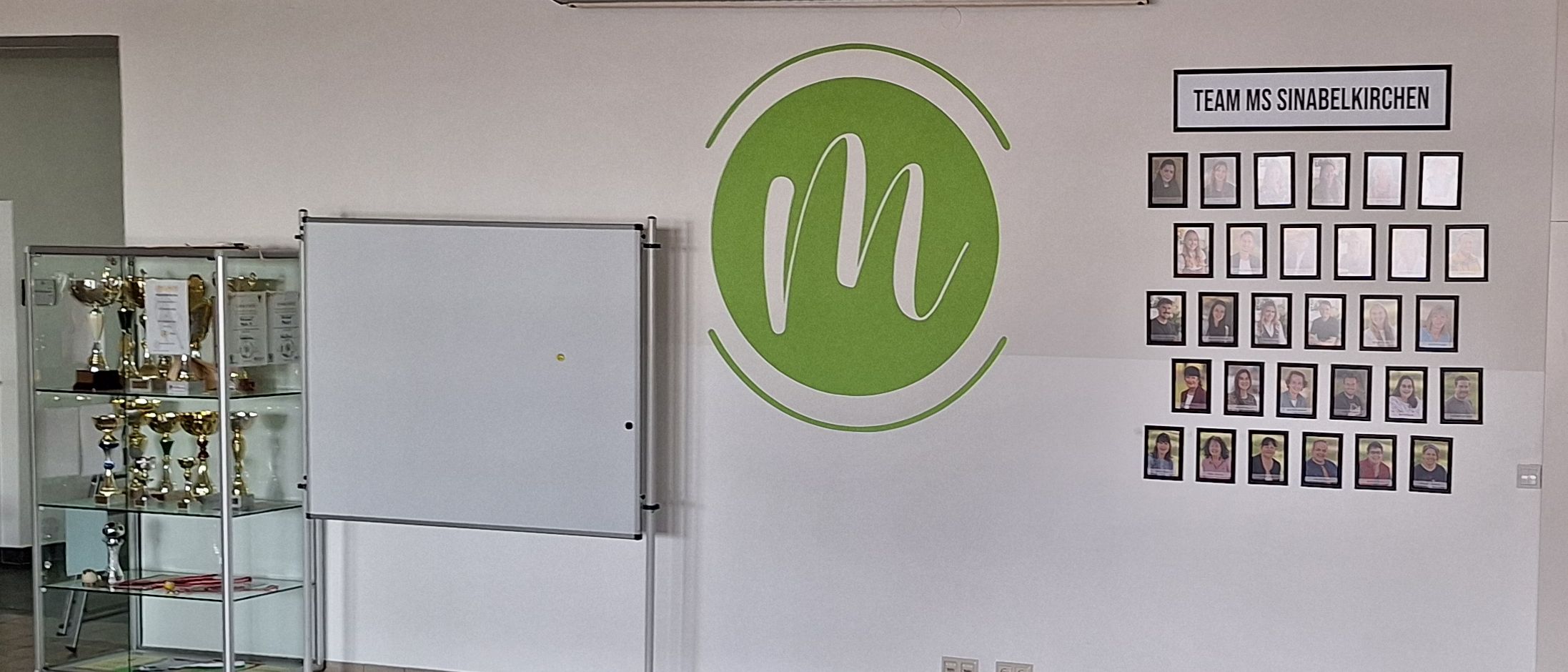 Ein leerer Raum mit einer weißen Wand, die ein grünes Logo und eine Pinnwand mit Fotos von Menschen trägt, die mit 'Team MS Sinabelkirchen' beschriftet sind.
