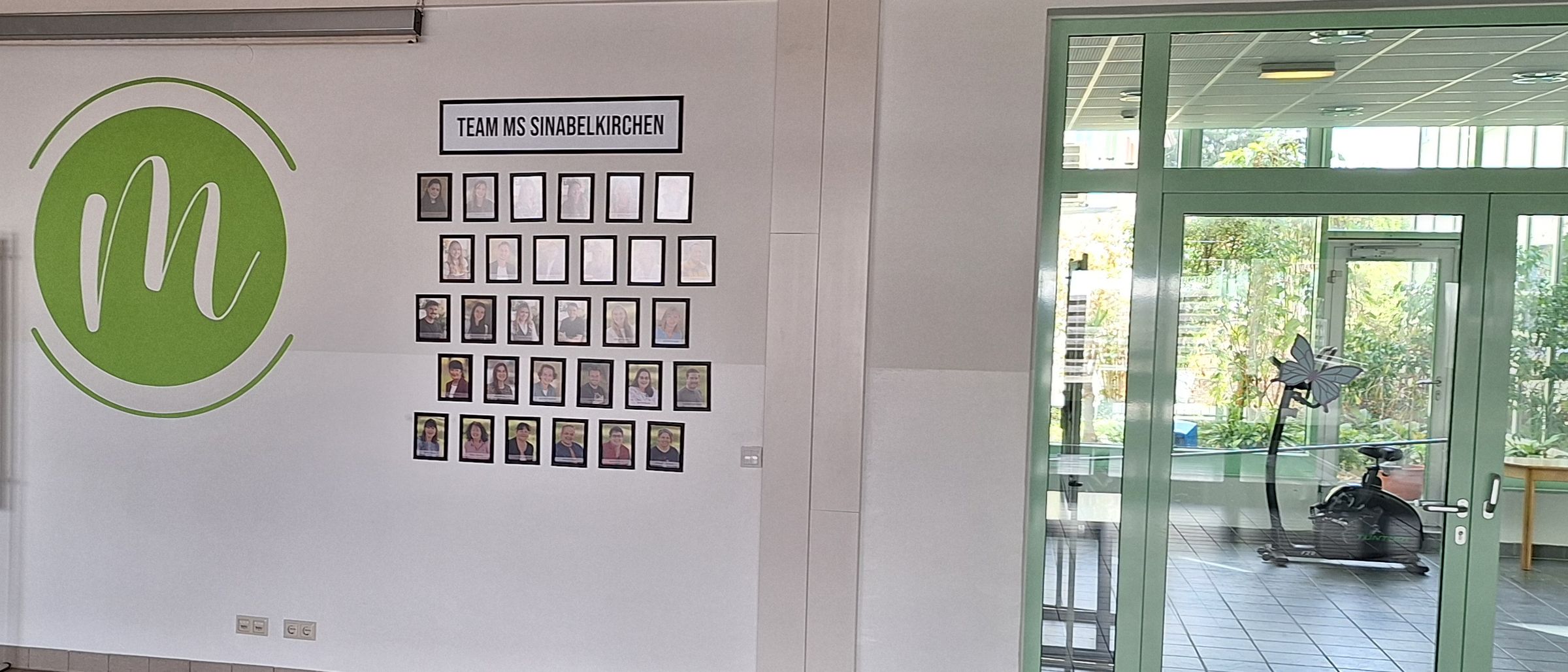 Ein leerer Raum mit einer weißen Wand, einer grünen Tür und einer Glastür. Es gibt eine Tafel, eine Glasvitrine und ein Logo an der Wand.