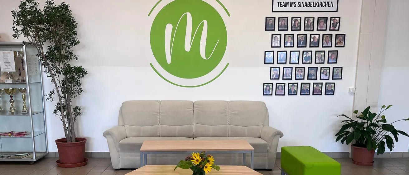 Ein Innenbereich mit einer Couch, einem Tisch und einem Topf. Die Wand hat einen grünen Kreis mit dem Buchstaben M und einem Teamnamen.
