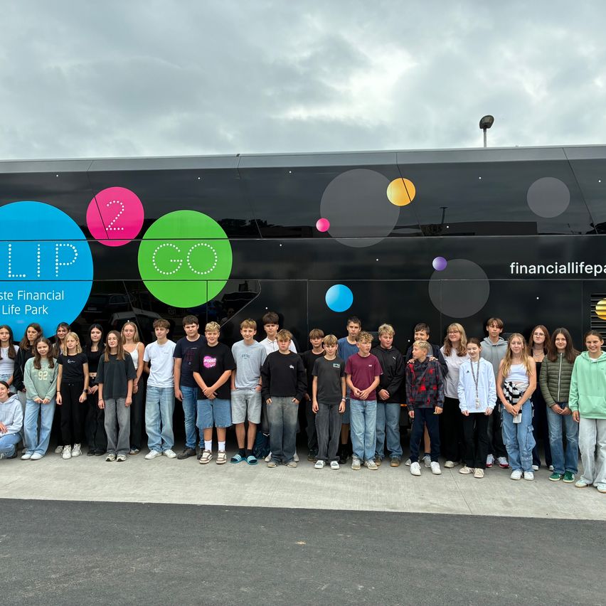 Eine Gruppe von Schülern steht vor einem Bus, der 'Flip Go' und 'financiallifepark' bewirbt. Die Schüler tragen Freizeitkleidung.