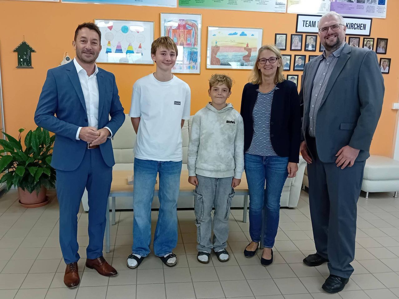 Fünf Personen posieren für ein Foto in einem Raum mit orangefarbenen Wänden und gerahmten Kunstwerken. Zwei Jungen stehen in der Mitte, mit zwei Erwachsenen, die auf beiden Seiten von ihnen stehen.