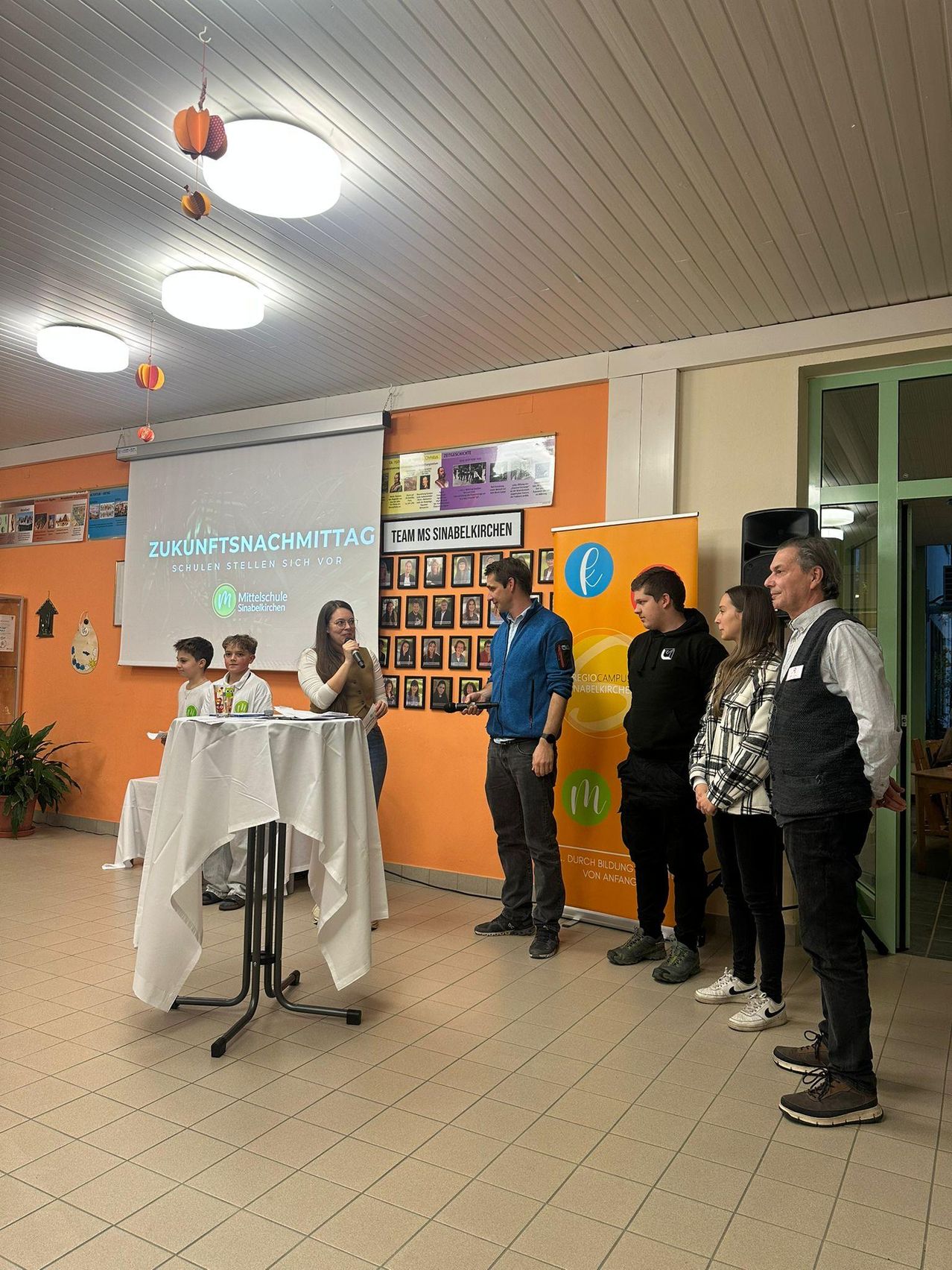 Mehrere Personen versammeln sich in einem Raum für eine Präsentation. Eine Frau spricht in ein Mikrofon an einem Tisch. Eine Projektionsleinwand zeigt den Text Zukunftsnachmittag. Der Raum hat eine orange Wand und eine Glastür.