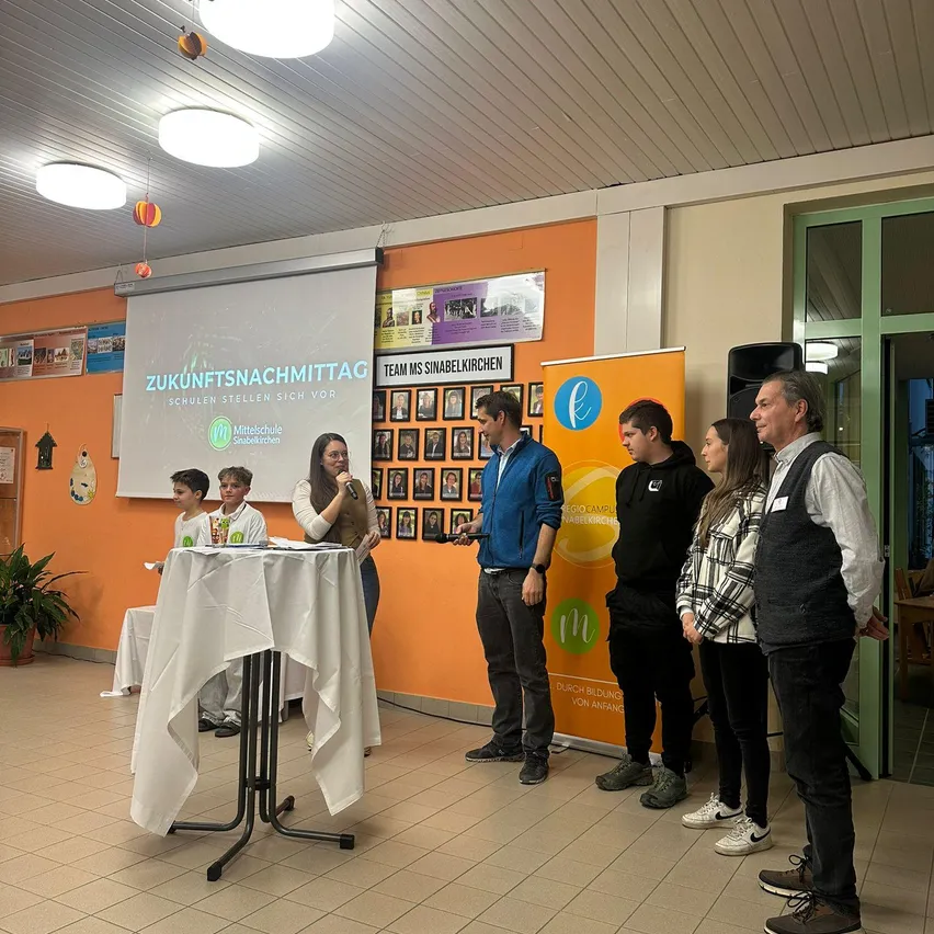 Mehrere Personen versammeln sich in einem Raum für eine Präsentation. Eine Frau spricht in ein Mikrofon an einem Tisch. Eine Projektionsleinwand zeigt den Text Zukunftsnachmittag. Der Raum hat eine orange Wand und eine Glastür.