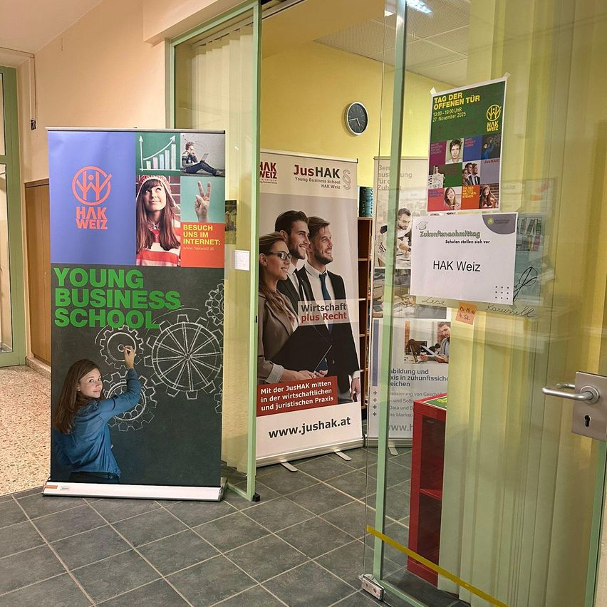 Ein Klassenzimmer zeigt Banner, die die Young Business School und JusHAK bewerben, mit einer Tafel und einem Mädchen, das zeichnet.