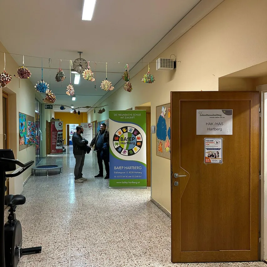 In einem Schulflur unterhalten sich zwei Personen in der Nähe eines Plakats, das eine freundliche Schule bewirbt. Ein Schild an der Tür lautet HAK/HAS Hartberg. Ein schwarzer Heimtrainer steht links.