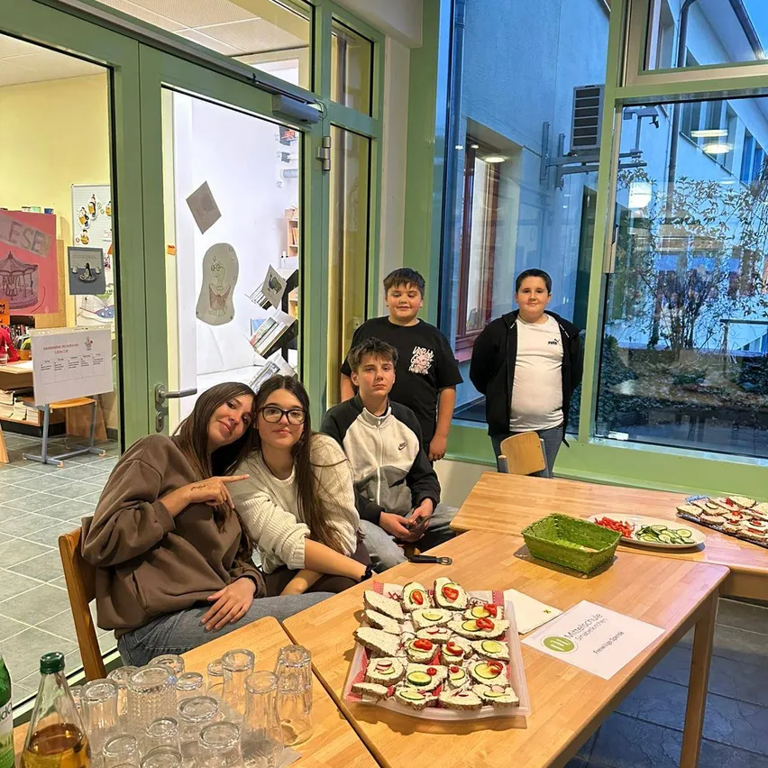 Eine Gruppe junger Menschen versammelt sich an einem Tisch mit Essen in einem Raum mit Glastüren.