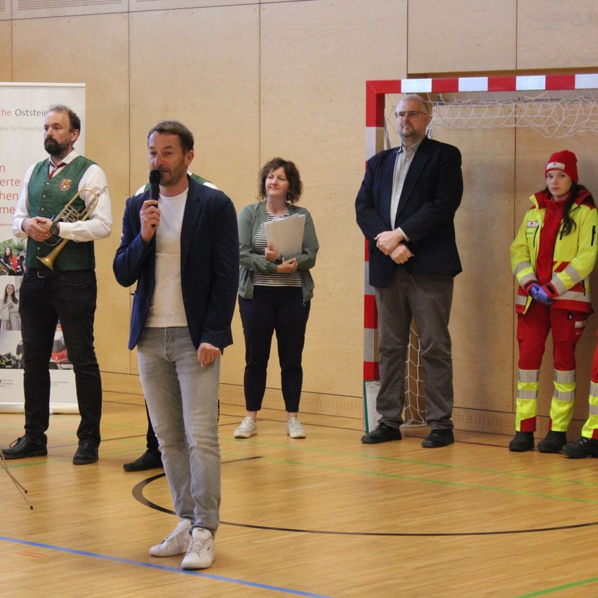 Fünf Personen stehen in einer Turnhalle. Ein Mann spricht in ein Mikrofon, während andere zusehen, darunter ein Mann mit Trompete. Eine Frau hält Papiere. Ein Fußballtor steht hinter ihnen.