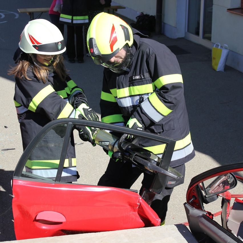 Zwei Feuerwehrleute in voller Ausrüstung verwenden ein Werkzeug, um eine beschädigte Autotür aufzuhebeln. Sie stehen neben dem Fahrzeug auf der Straße.