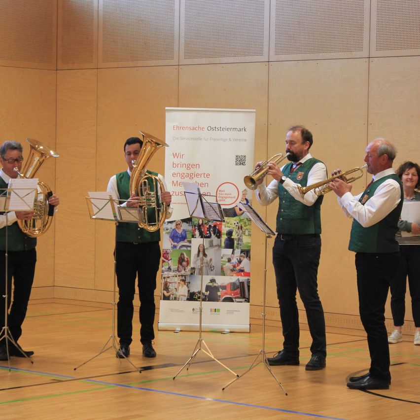 Fünf Musiker in grünen Westen und schwarzen Hosen spielen Blechblasinstrumente auf einer Bühne vor einem Banner. Zwei Personen stehen hinter ihnen und beobachten.