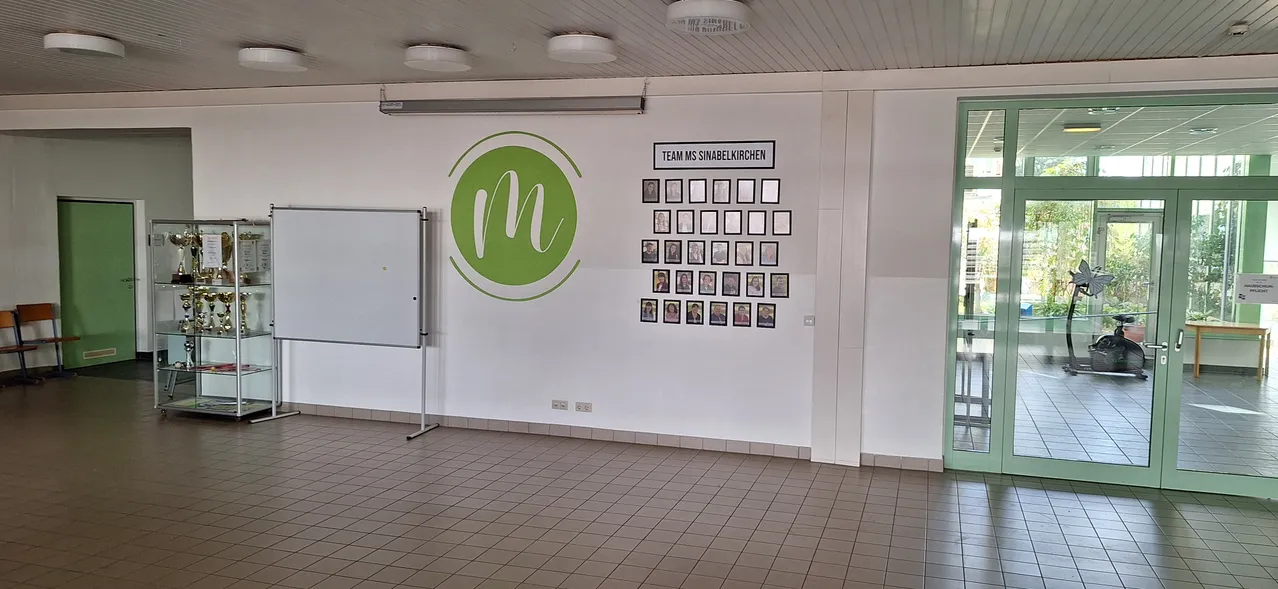 Ein leerer Raum mit einer weißen Wand, die ein grünes Logo und eine Pinnwand mit Fotos von Menschen zeigt.