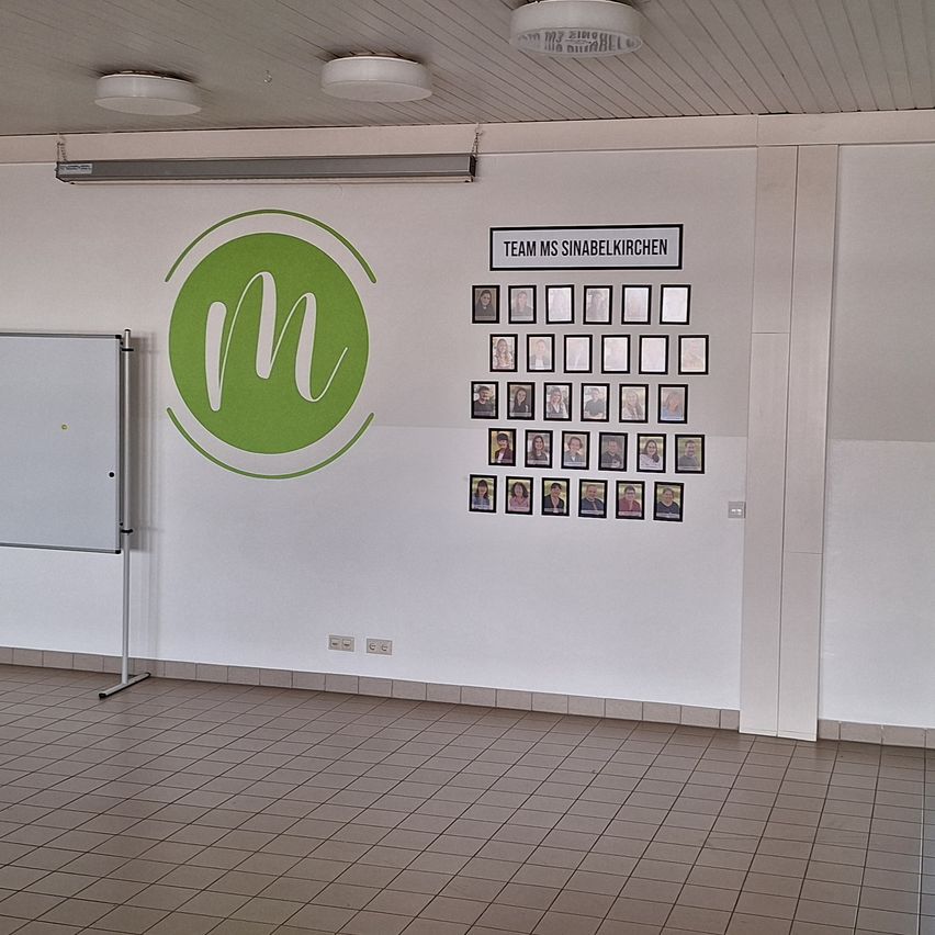 Ein leerer Raum mit einer weißen Wand, die ein grünes Logo und eine Pinnwand mit Fotos von Menschen zeigt.