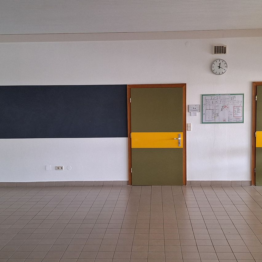 Ein leerer Raum mit gefliestem Boden, einer schwarzen und gelben Wand, einer grünen Tür und einer weißen Wand mit einer Uhr und einer Pinnwand.