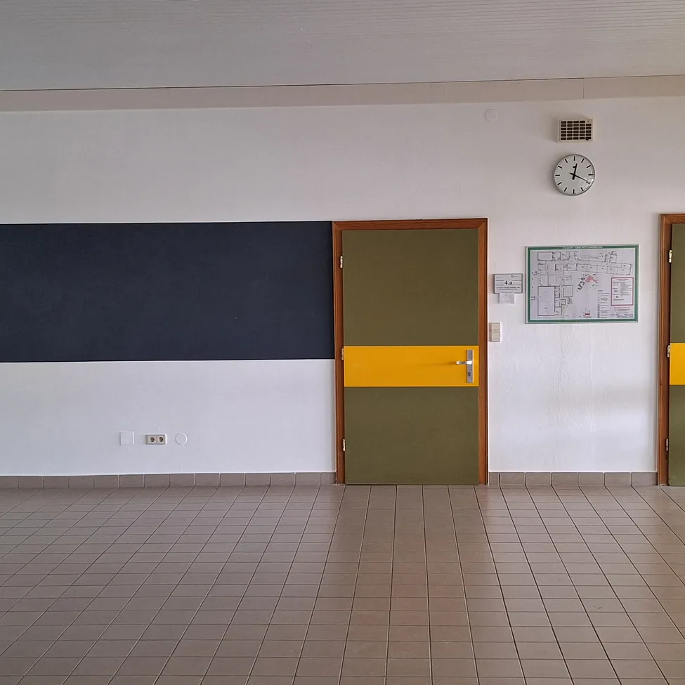 Ein leerer Raum mit gefliestem Boden, einer schwarzen und gelben Wand, einer grünen Tür und einer weißen Wand mit einer Uhr und einer Pinnwand.