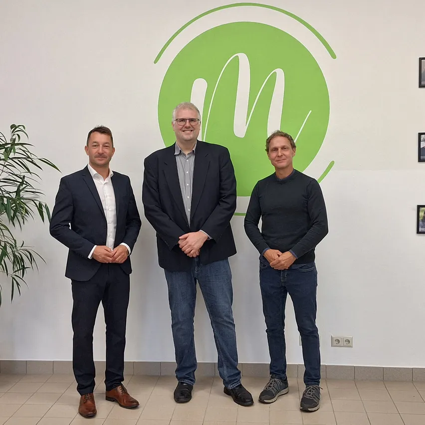 Drei Männer stehen zusammen vor einer weißen Wand mit einem grünen kreisförmigen Logo. Der Mann auf der linken Seite trägt einen dunklen Anzug, der Mann in der Mitte trägt einen dunklen Blazer über einem grauen Hemd und der Mann auf der rechten Seite trägt ein dunkles Langarmhemd. Sie haben alle ihre Hände in den Taschen.