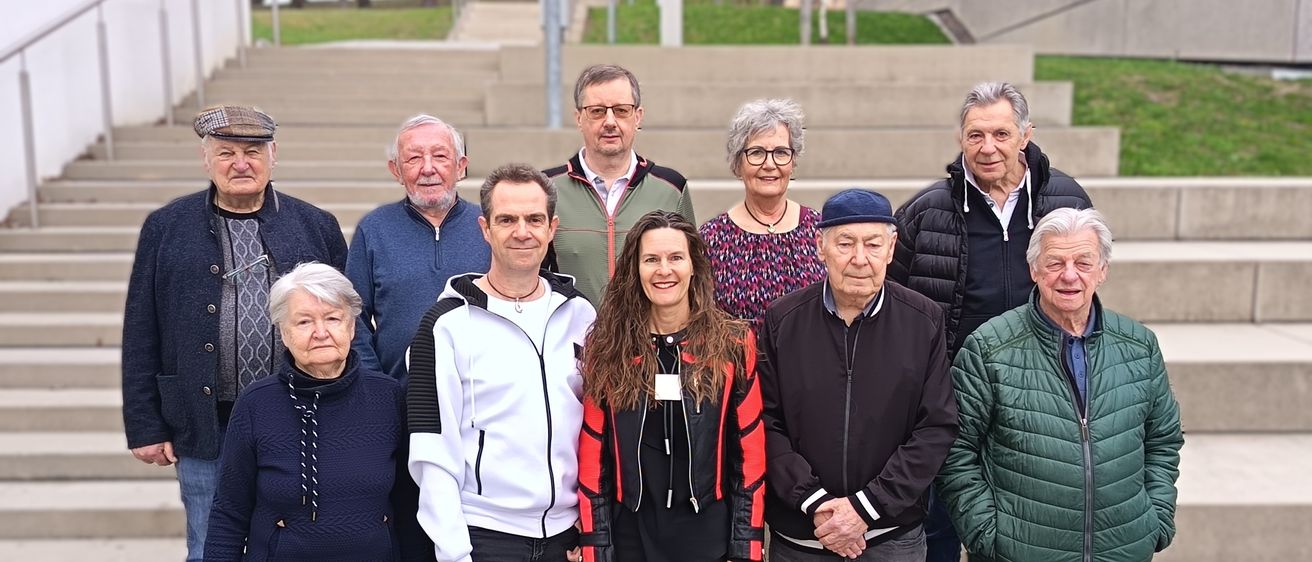 Eine Gruppe älterer Menschen posiert für ein Foto auf Betonstufen mit Grün. Sie tragen verschiedene Jacken und Accessoires. Die Frau in der Mitte trägt eine rot-schwarze Jacke.
