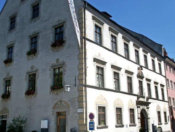Ein großes weißes Gebäude mit mehreren Fenstern und Topfpflanzen davor, ein Fahrrad geparkt in der Nähe und ein Schild, das auf einen Fußgängerüberweg hinweist.