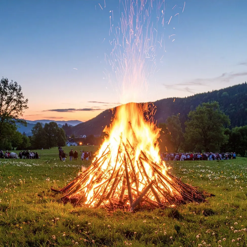 Ein großes Lagerfeuer brennt hell in einer grasigen Wiese, mit Menschen, die drum herum stehen und sitzen und zuschauen. Bäume und Berge sind im Hintergrund.