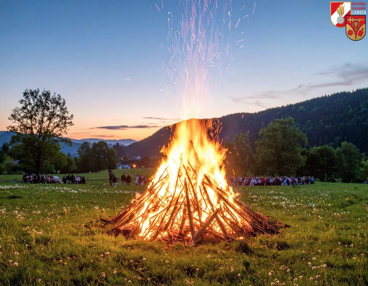 Ein großes Lagerfeuer brennt hell in einer grasigen Wiese, mit Menschen, die drum herum stehen und sitzen und zuschauen. Bäume und Berge sind im Hintergrund.