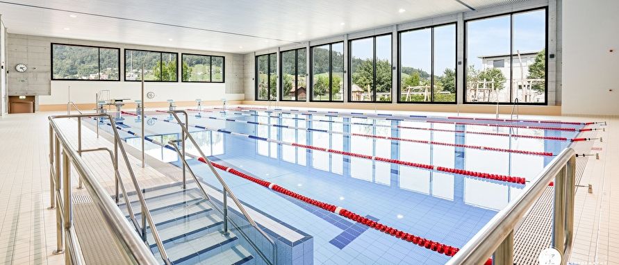 An indoor swimming pool with multiple lanes, red lane ropes, and large glass windows overlooking a landscape.