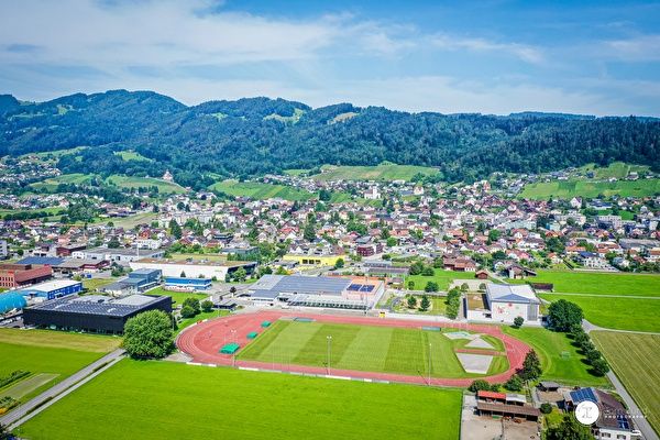 Eine Luftaufnahme einer Stadt mit vielen Gebäuden, einem Sportplatz und Bergen im Hintergrund.