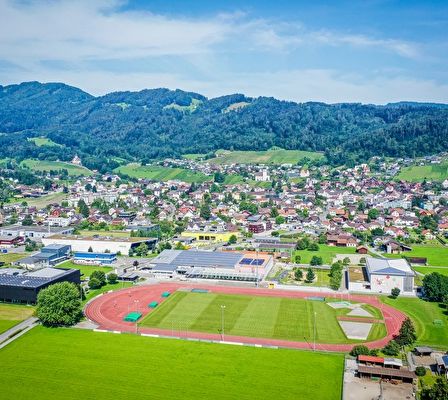Eine Luftaufnahme einer Stadt mit vielen Gebäuden, einem Sportplatz und Bergen im Hintergrund.