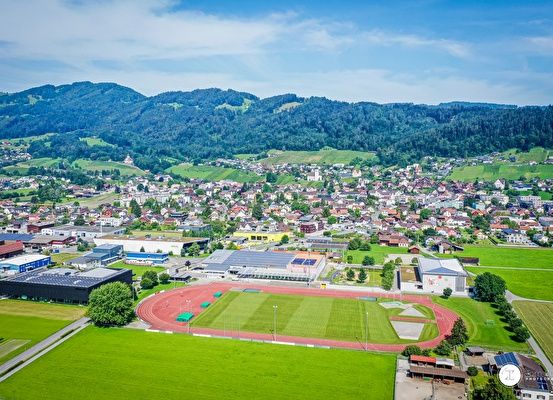 An aerial view of a town with many buildings, a sports field, and mountains in the background.