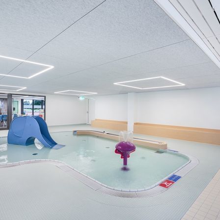 Ein Hallenbad mit einer Rutsche, einem Springbrunnen und weißen Fliesen auf dem Boden. Es gibt einen Raum mit Glaswänden und einem Tisch mit Stühlen.